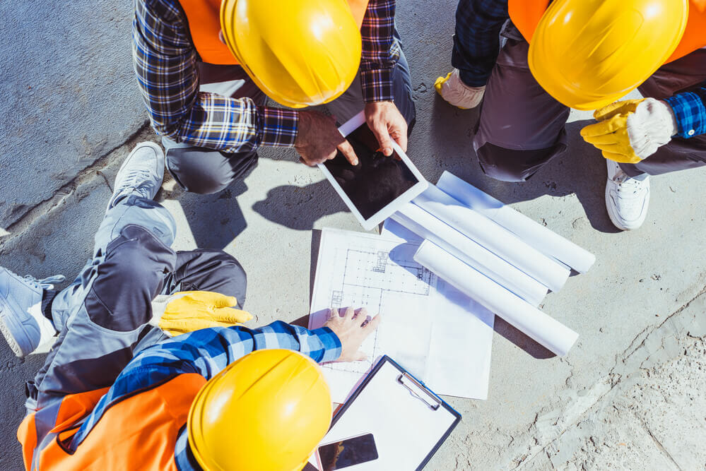 Three workers huddled around blueprint papers and an iPad in preparation for roof construction