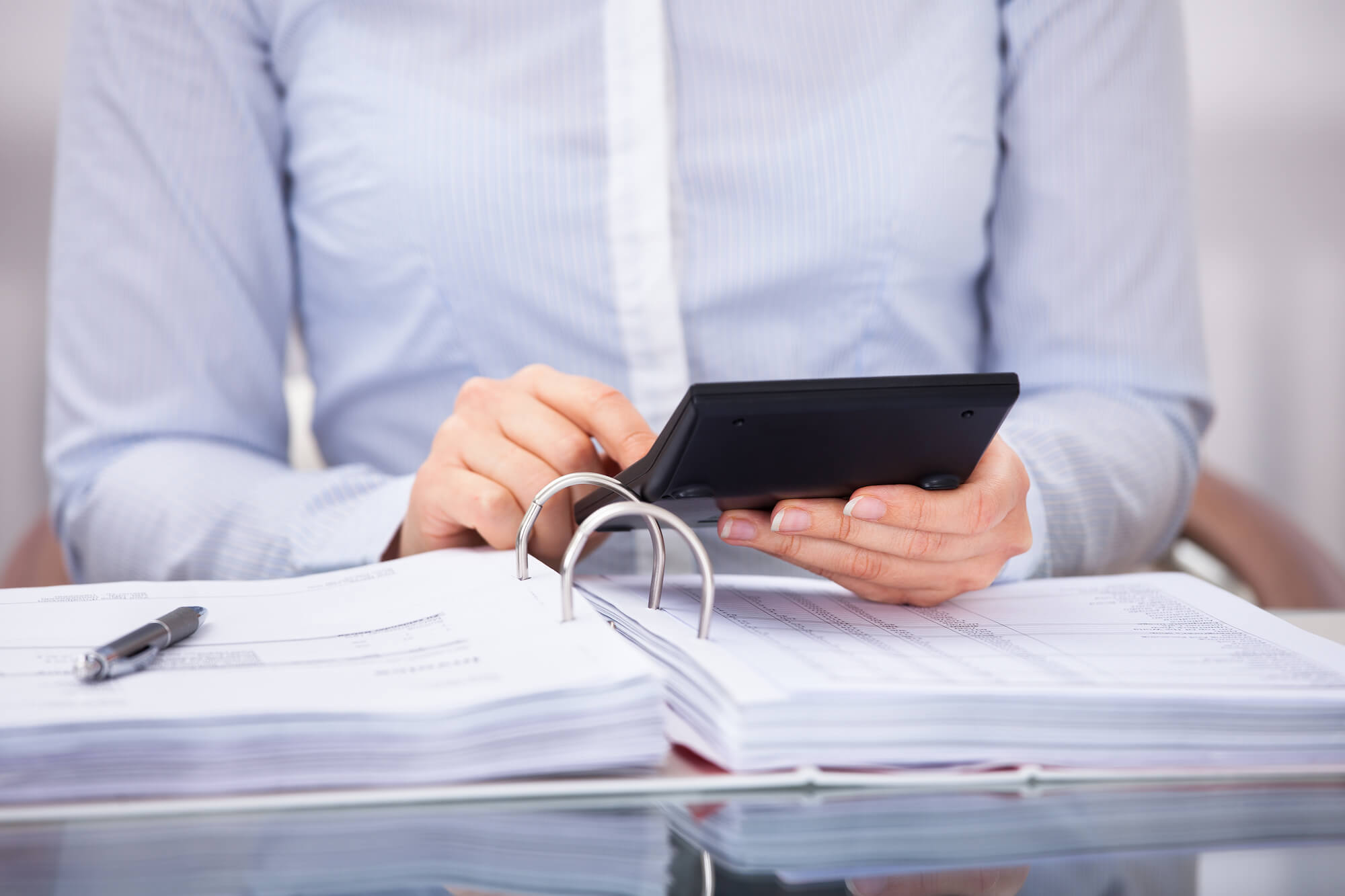 women in blue dress shirt using a calculator in with a binder and pen on a desk