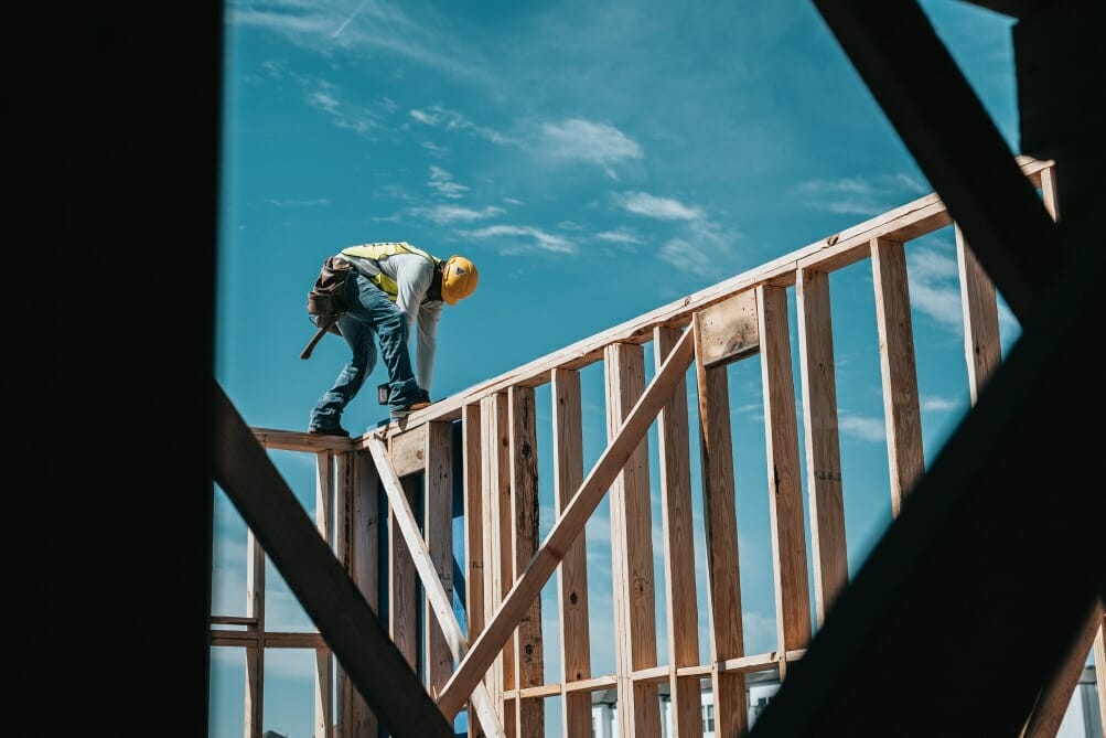 Construction worker walking on the frame of a building with a hardhat on.